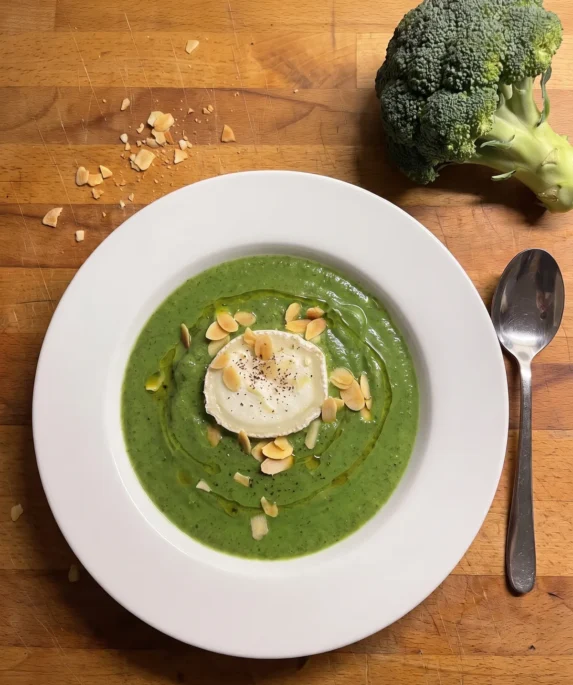 Gordon Ramsay broccoli soup: bright green blended broccoli soup beside a slice of goat's cheese, with toasted sliced almonds and olive oil in a shallow bowl