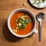Gordon Ramsay roasted red pepper and tomato soup: chilled orange-red soup in a white soup cup topped with diced cucumber, a basil sprig and olive oil