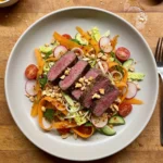 Gordon Ramsay spicy beef salad: thick slices of medium-rare sirloin steak over carrot ribbons, radishes, cherry tomatoes, cucumber, baby gem and mint with crushed peanuts