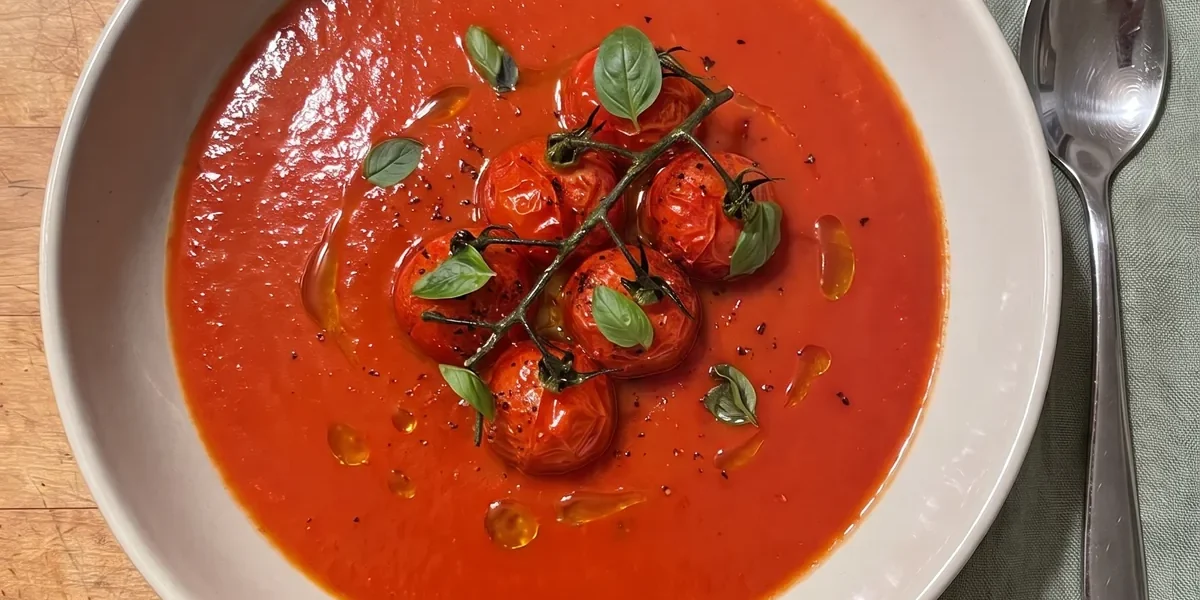 Gordon Ramsay tomato basil soup: smooth red tomato soup in a wide bowl topped with a cluster of pan-fried cherry tomatoes on the vine and small basil leaves