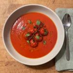 Gordon Ramsay tomato basil soup: smooth red tomato soup in a wide bowl topped with a cluster of pan-fried cherry tomatoes on the vine and small basil leaves