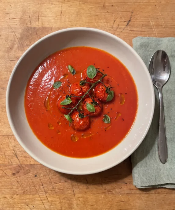 Gordon Ramsay tomato basil soup: smooth red tomato soup in a wide bowl topped with a cluster of pan-fried cherry tomatoes on the vine and small basil leaves
