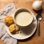 Gordon Ramsay white onion soup: a handled bowl of smooth pale onion and cheddar soup with two warm scones on the plate
